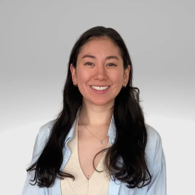 A young woman with long dark hair, smiling, wearing a light blue shirt over a beige sweater, standing against a neutral background.
