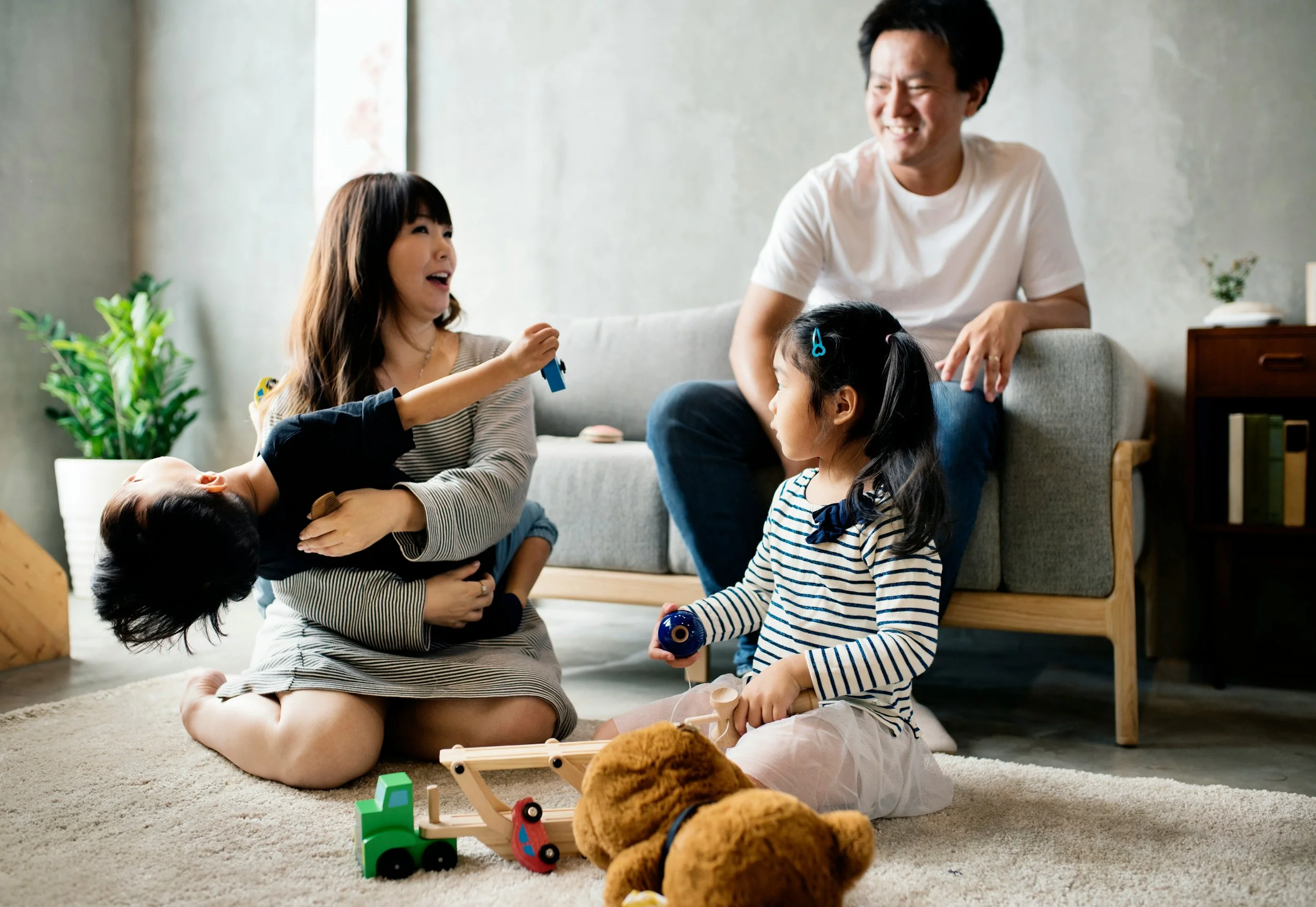 A family of four, including a mother, father, and two young daughters, playing together in a living room. The mother is holding one of the daughters who is reaching out with a toy. The father is sitting on a sofa smiling at the children. The other daughter is sitting on the floor with toys around her.