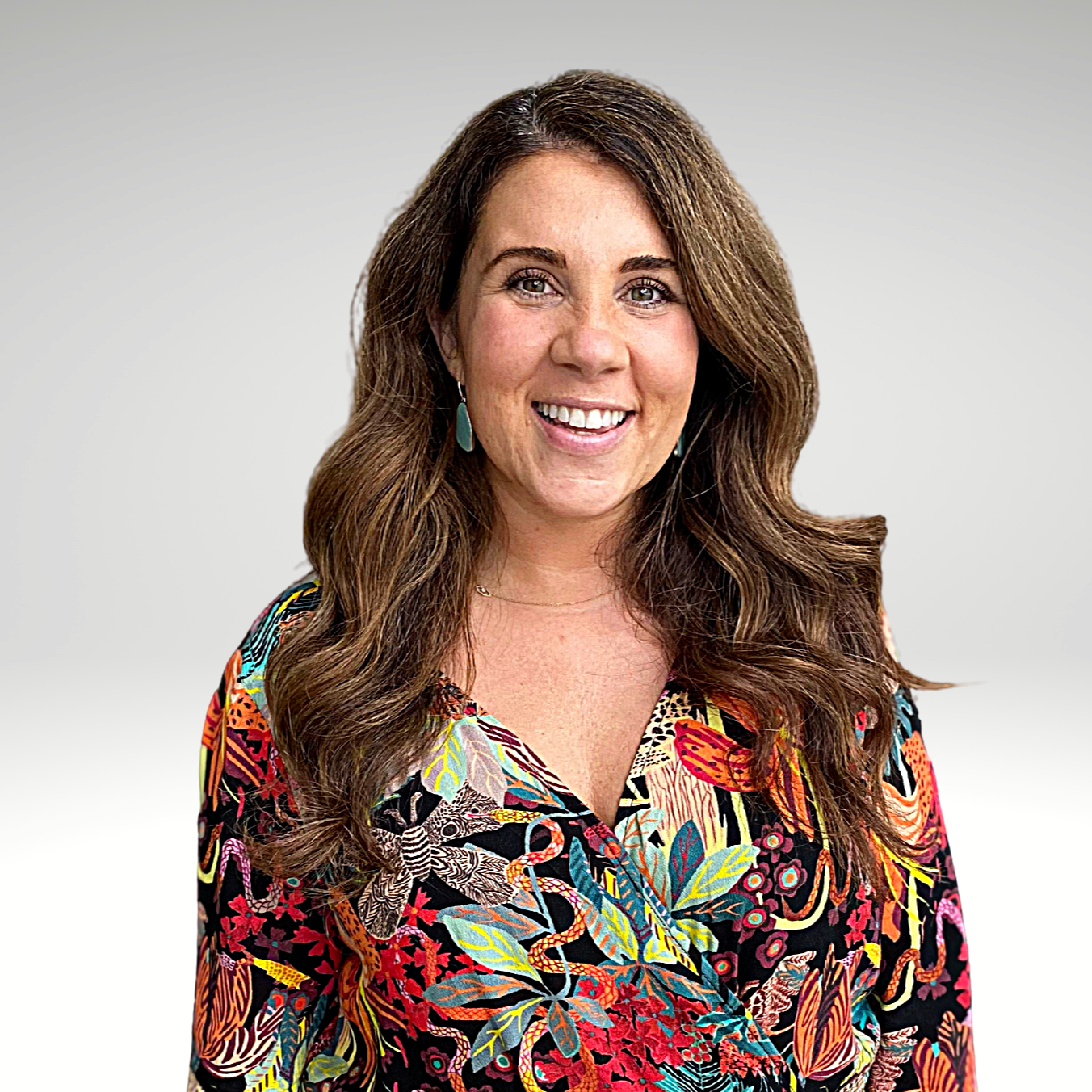 A woman with wavy brown hair smiling, wearing a colorful floral-patterned top and turquoise earrings.