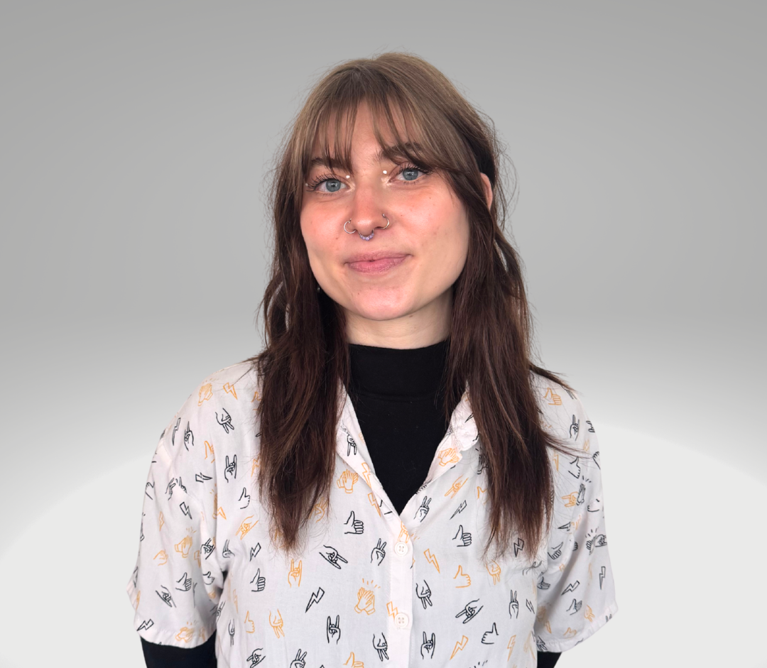 A woman with brown hair, blue eyes, and a septum piercing stands against a plain gray background, wearing a white button-up shirt with black and orange hand gesture icons over a black long-sleeve shirt.