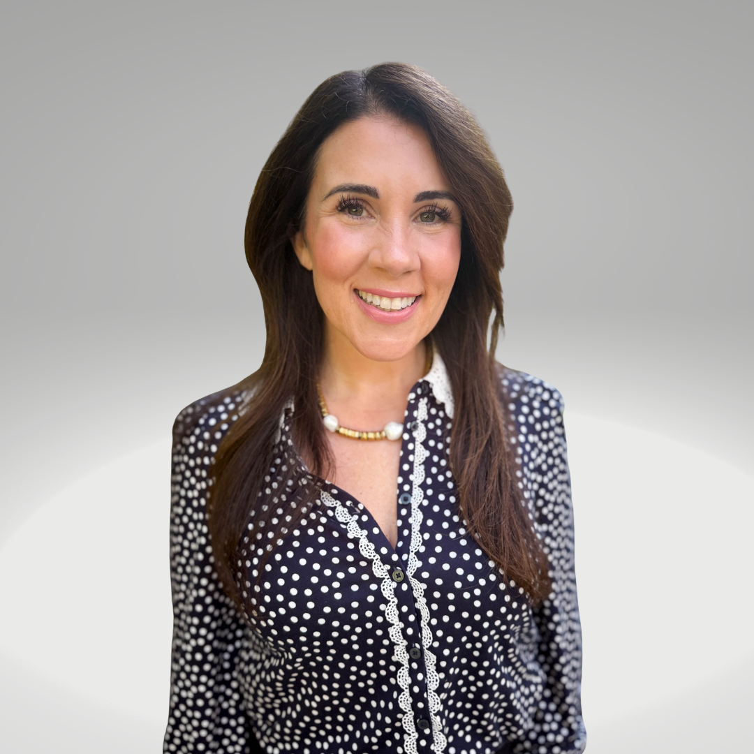 A woman with long, wavy brown hair, smiling and wearing a colorful floral blouse with bright earrings, against a plain gray background.