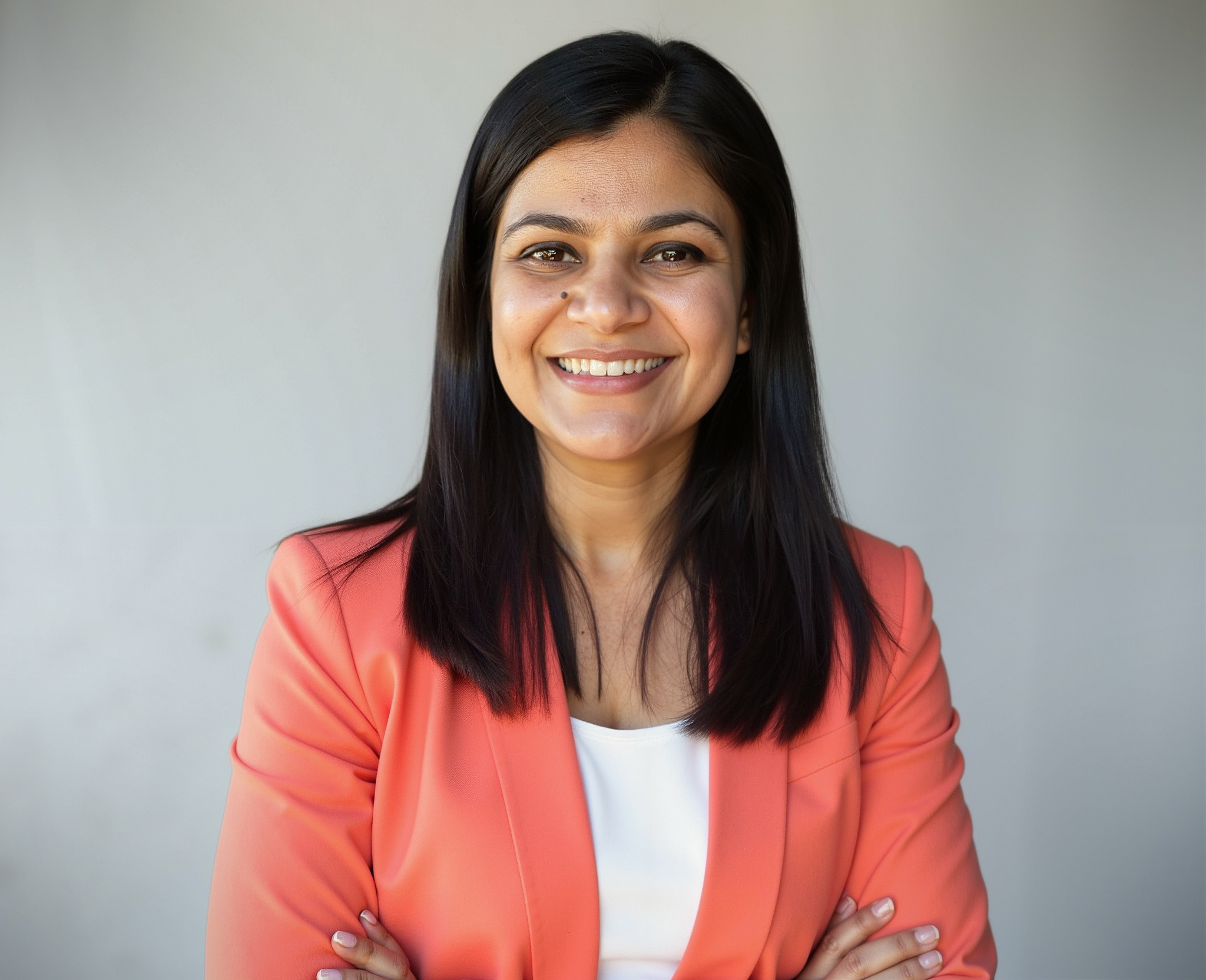 A woman with long dark hair, wearing a coral blazer and white top, smiling with arms crossed against a light gray background.