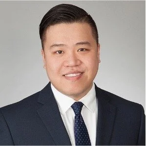 Professional headshot of a man wearing a suit and tie, smiling against a gray background.