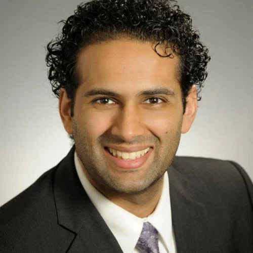 Headshot of a smiling man with curly dark hair, wearing a black suit jacket, white shirt, and a purple tie.
