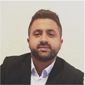 A man with dark hair and a beard, wearing a black blazer and white shirt, posing against a plain background.