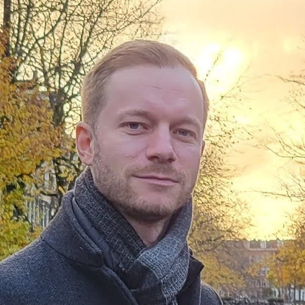 A man with short blonde hair and a beard posing outdoors during sunrise or sunset, with trees and buildings in the background.