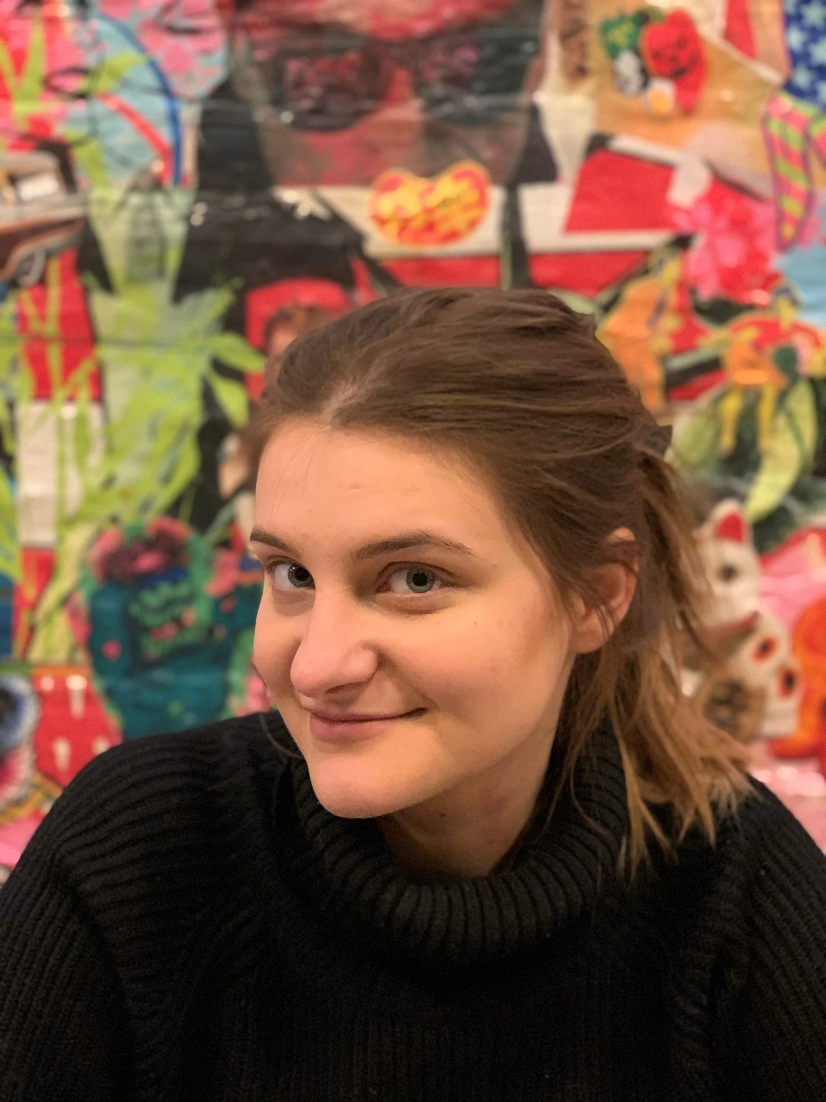 A woman with shoulder-length wavy light brown hair and blue eyes, smiling in front of a colorful, abstract background.