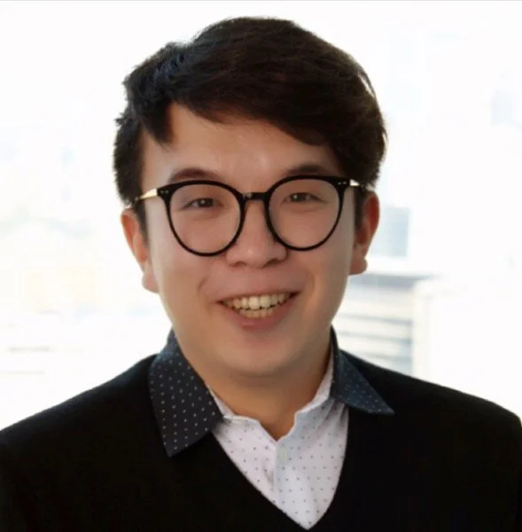 A young man with short dark hair, wearing black-rimmed glasses, a white shirt with small dots, and a dark blazer, smiling at the camera with a bright window in the background.