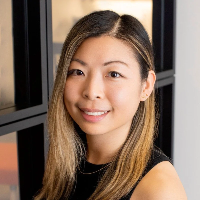 A young woman with long, straight hair smiling at the camera, standing near black shelves.