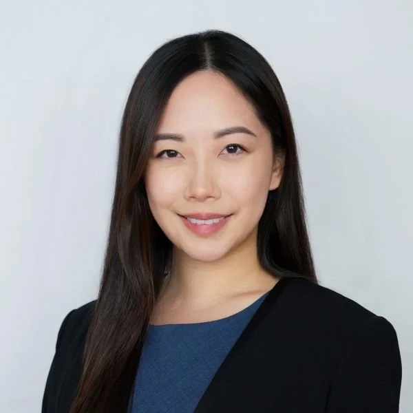 A young Asian woman with long dark hair, smiling, wearing a black blazer and blue top, against a light grey background.