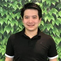 A young man smiling, wearing a black polo shirt with white trim, standing in front of a green leafy background.