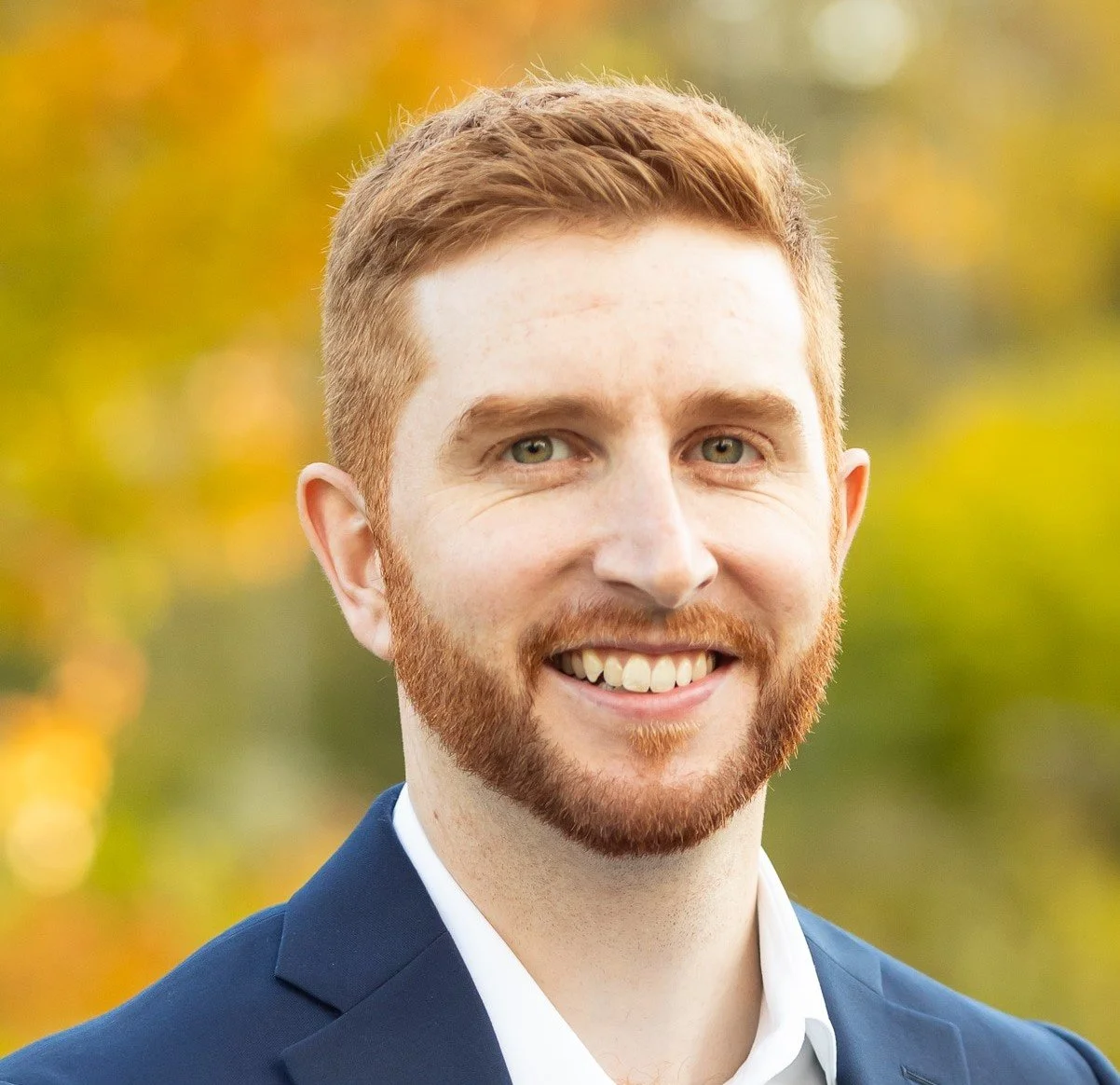 A young man with red hair, a beard, and blue eyes is smiling and wearing a navy blue suit with a white shirt. The background is blurred with autumn colors.