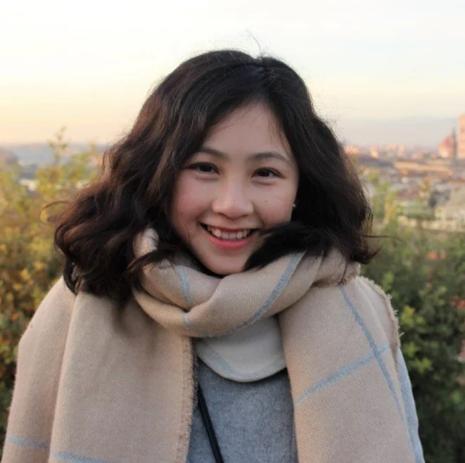 Smiling woman with curly hair wearing a beige and gray scarf outdoors during sunset with cityscape in the background.