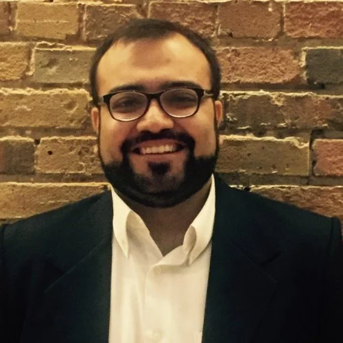 A man with glasses and a beard smiling in front of a brick wall, wearing a black suit and white shirt.