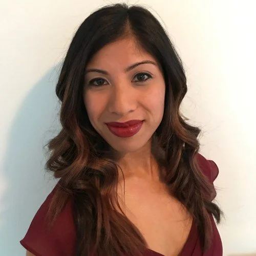 A woman with long, wavy hair and red lipstick smiling in front of a white wall.