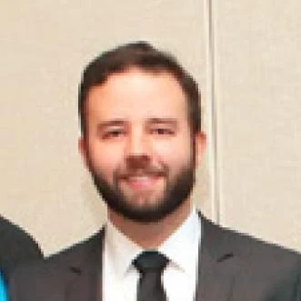 A man with a beard wearing a suit and tie, smiling, with a plain background.