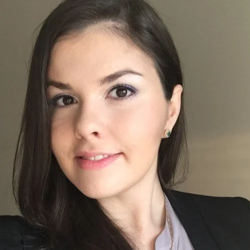 Close-up of a young woman with long brown hair, light makeup, wearing a black blazer and a white shirt, smiling slightly at the camera.