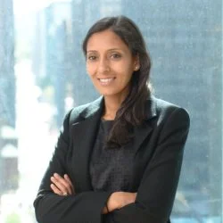 A woman smiling with crossed arms, wearing a black blazer and dark top, standing in front of a glass window in an office setting.