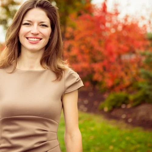 A smiling woman with shoulder-length brown hair standing outdoors in front of autumn-colored trees and greenery.