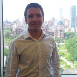A man standing indoors with a cityscape background, wearing a light-colored checkered shirt and smiling.