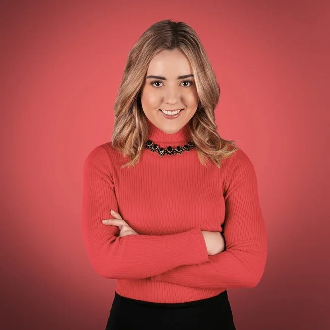 A young woman with blonde, shoulder-length hair smiling, wearing a red ribbed turtleneck sweater and a black necklace, against a pink background.