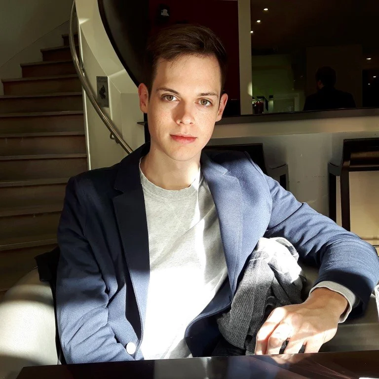A young man sitting at a table in a restaurant or café, wearing a navy blazer over a light-colored shirt, with short dark hair and a neutral expression.