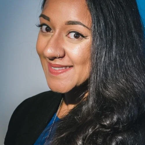 Close-up of a woman with dark hair, a nose piercing, and light makeup, smiling slightly, wearing a black top and a blue shirt.