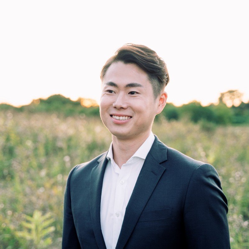 A smiling young man in a suit standing outdoors in a field during sunset.