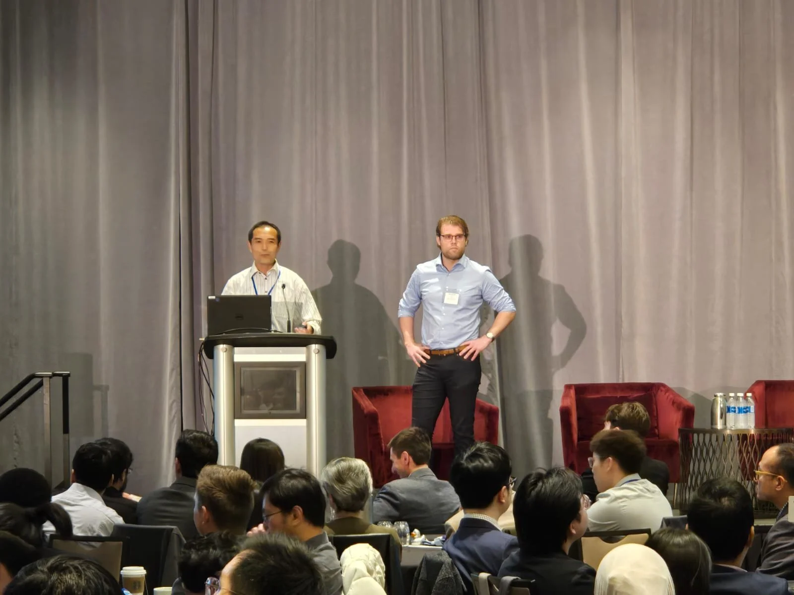 Two speakers on stage at a conference, one standing at a podium with a laptop, the other standing with hands on hips, audience seated watching.
