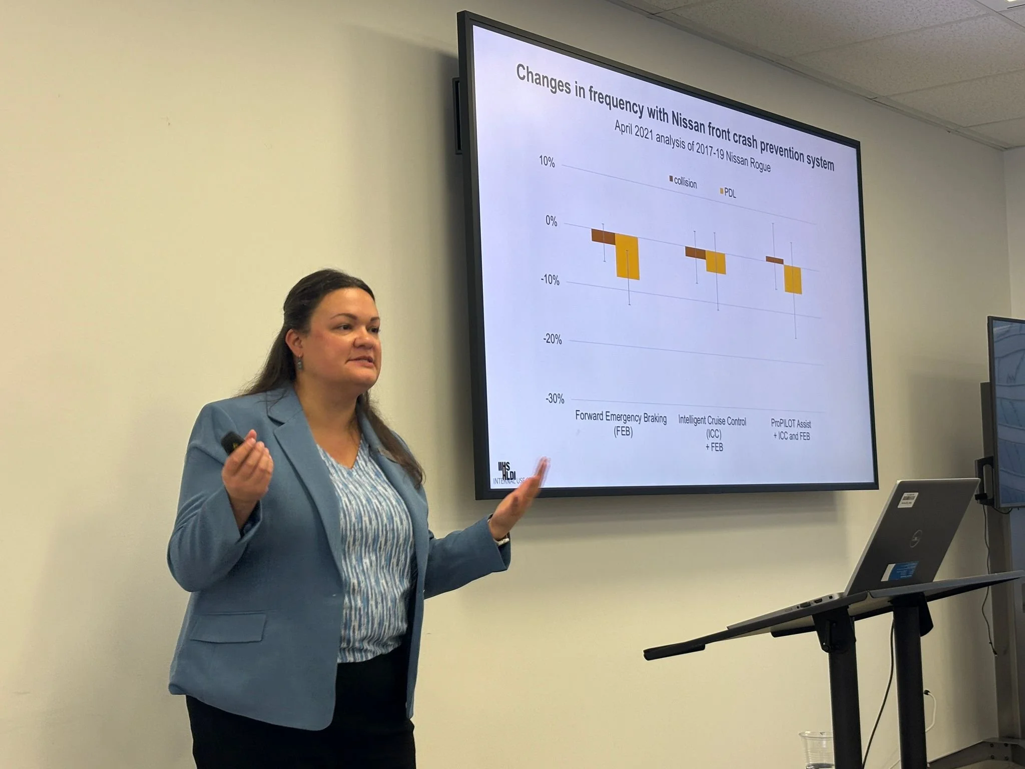 A woman giving a presentation in front of a large screen displaying a graph about Nissan crash prevention systems, with a laptop on a stand nearby.