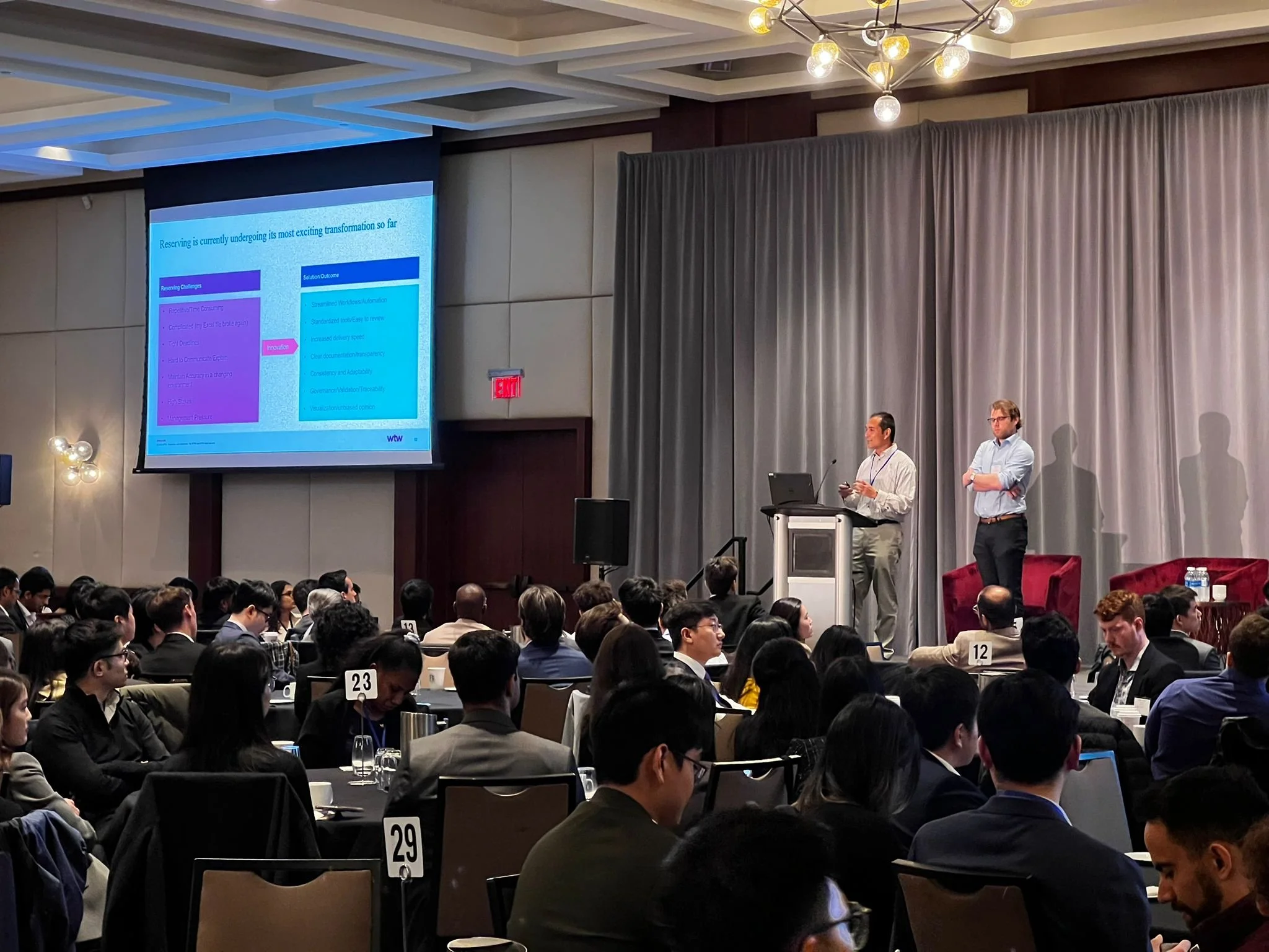 Two presenters standing on stage near a large screen at a conference, with an audience seated at round tables. The slide on the screen shows a presentation about transformation challenges and solutions.