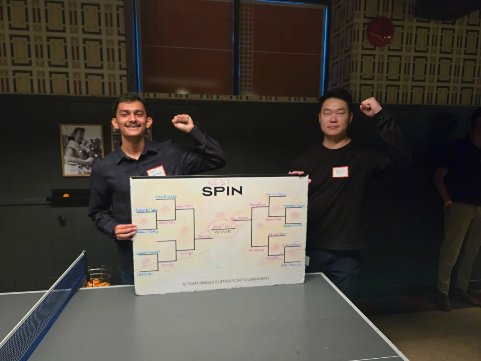 Two young men standing behind a ping pong tournament bracket, smiling, with fists raised. The man on the left has a mustache and is wearing a dark shirt, the man on the right is wearing a black shirt. The background includes framed photos and a patterned wall.
