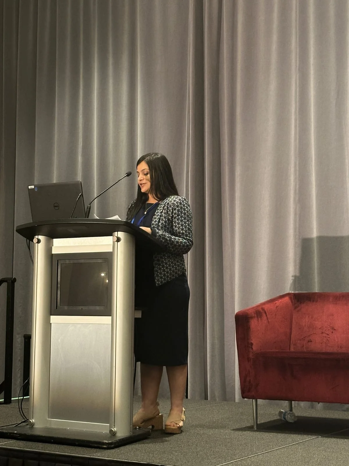 A woman with black hair, wearing a black dress and a patterned blazer, stands behind a podium with a laptop, speaking into a microphone in front of gray curtains, with a red chair to her right.