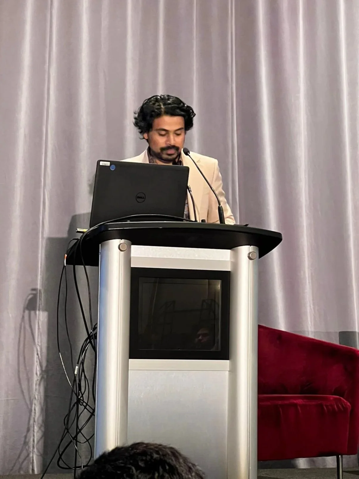 Man with curly hair and beard wearing a beige blazer speaking at a podium with a laptop, in front of gray curtain backdrop.