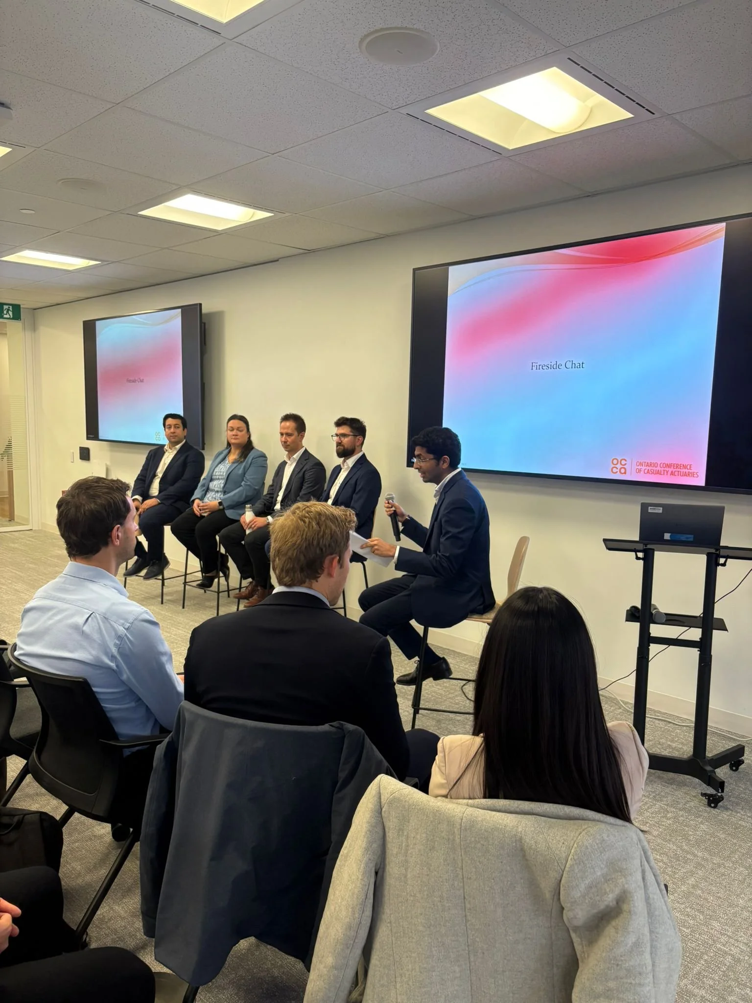 A panel discussion at a conference with five speakers seated in front of an audience. One speaker is holding a microphone and speaking, with a large screen behind displaying 'Fireside Chat' and the Ontario Conference of Casualty Actuaries logo.