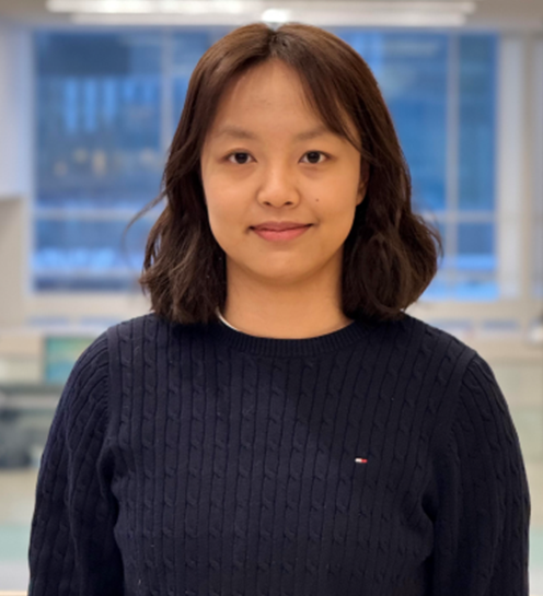 A woman with shoulder-length dark hair, wearing a navy blue sweater, standing indoors with a blurred cityscape background visible through large windows.
