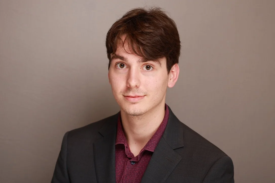 A young man with brown hair, wearing a dark suit jacket and a maroon shirt, looking at the camera with a neutral expression, against a plain gray background.