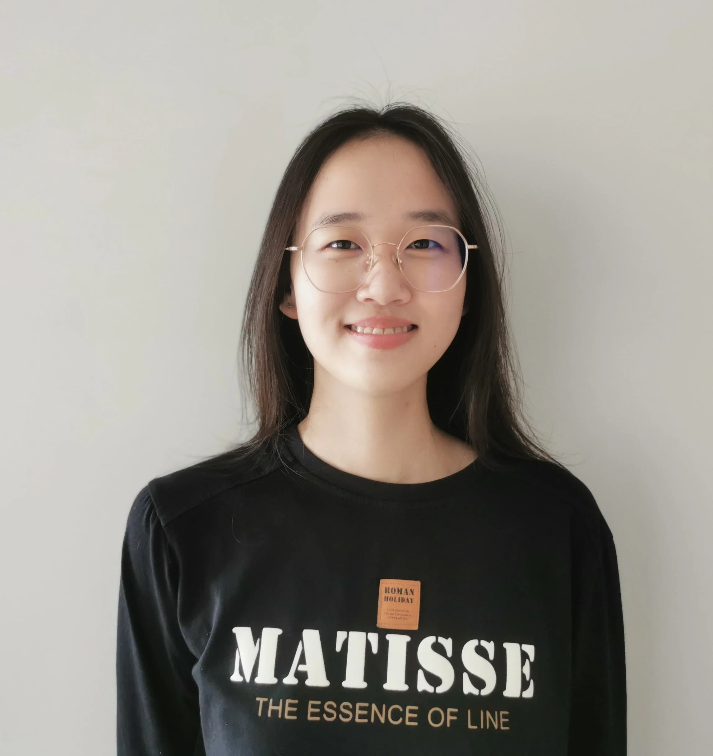 A young woman with long dark hair and glasses smiling in front of a plain white wall, wearing a black shirt with text.
