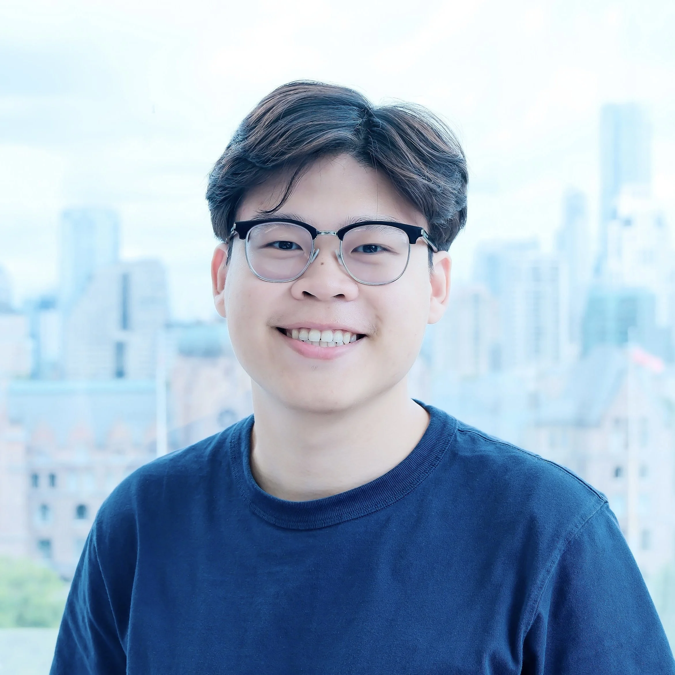 Young man smiling with glasses, wearing a dark t-shirt, in front of a cityscape with tall buildings.