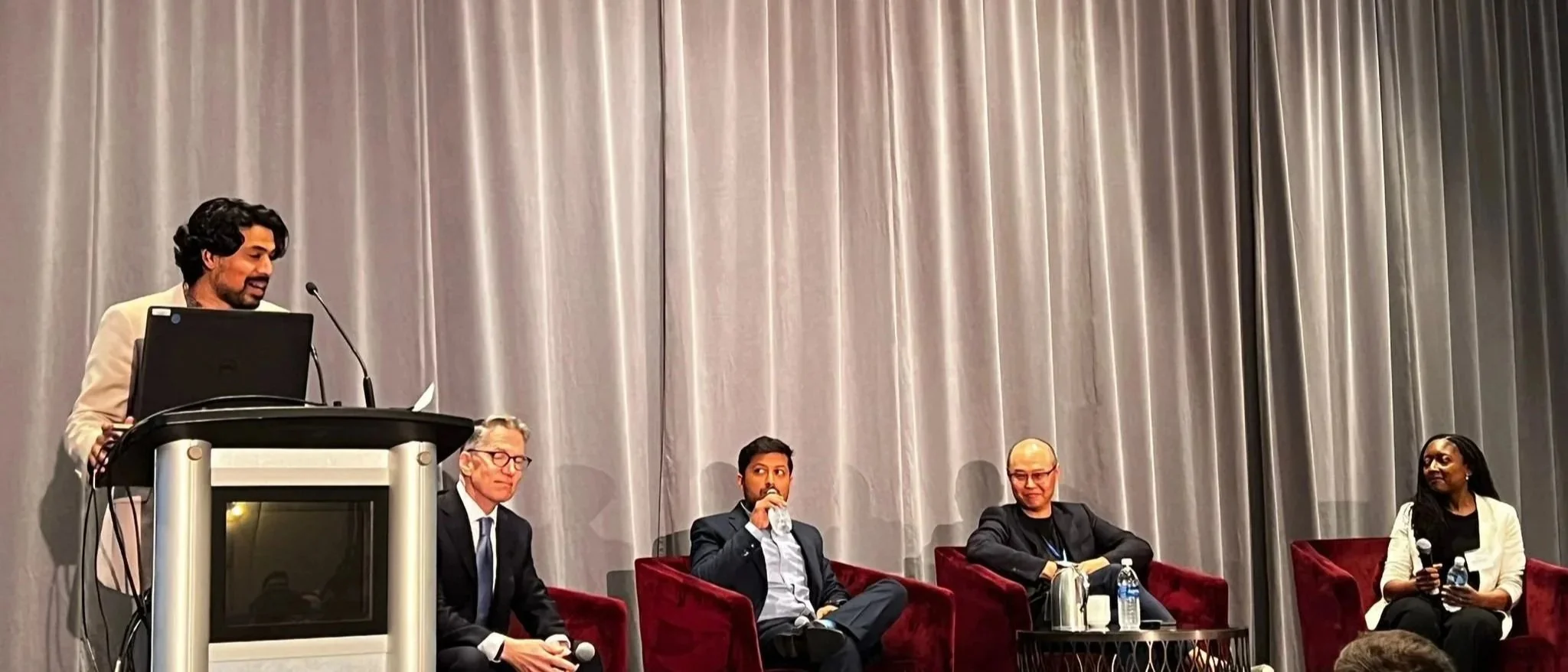 A panel discussion with five individuals, one person standing at a podium with a microphone and four seated on velvet chairs, with a gray curtain backdrop.