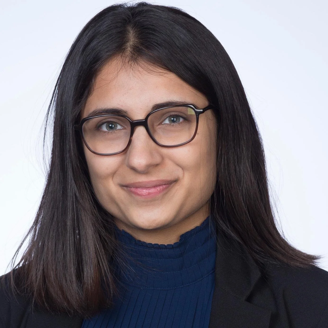 A woman with shoulder-length dark brown hair, wearing black-rimmed glasses, a blue turtleneck, and a black blazer, smiling, against a plain white background.