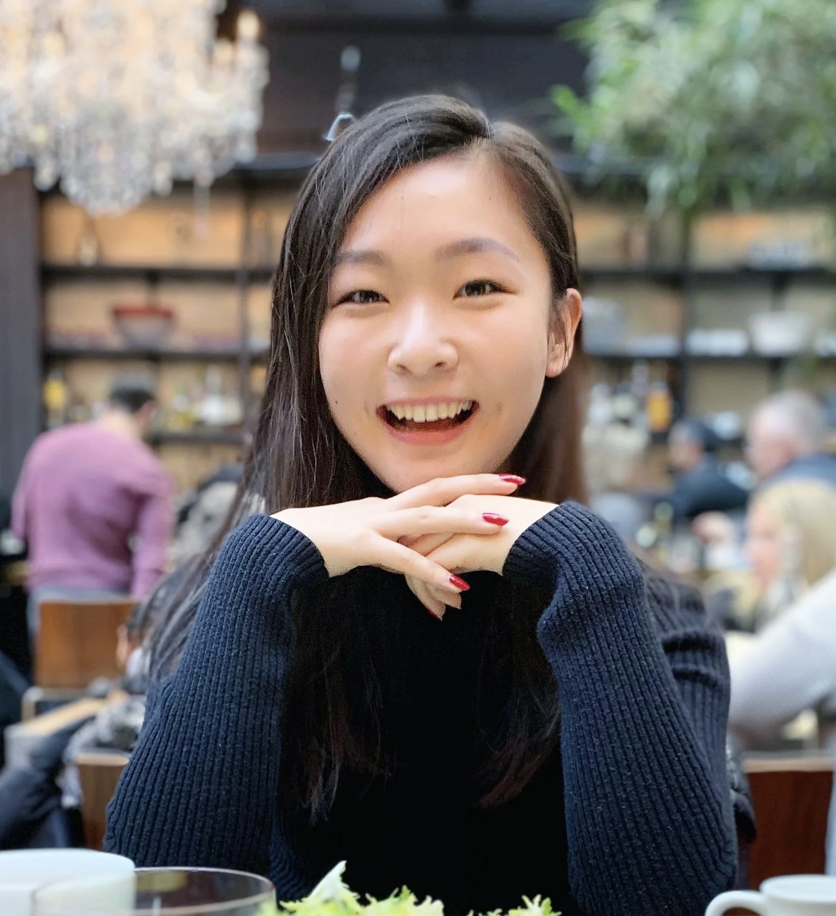 A young woman with long dark hair smiling and resting her chin on her clasped hands at a restaurant table.