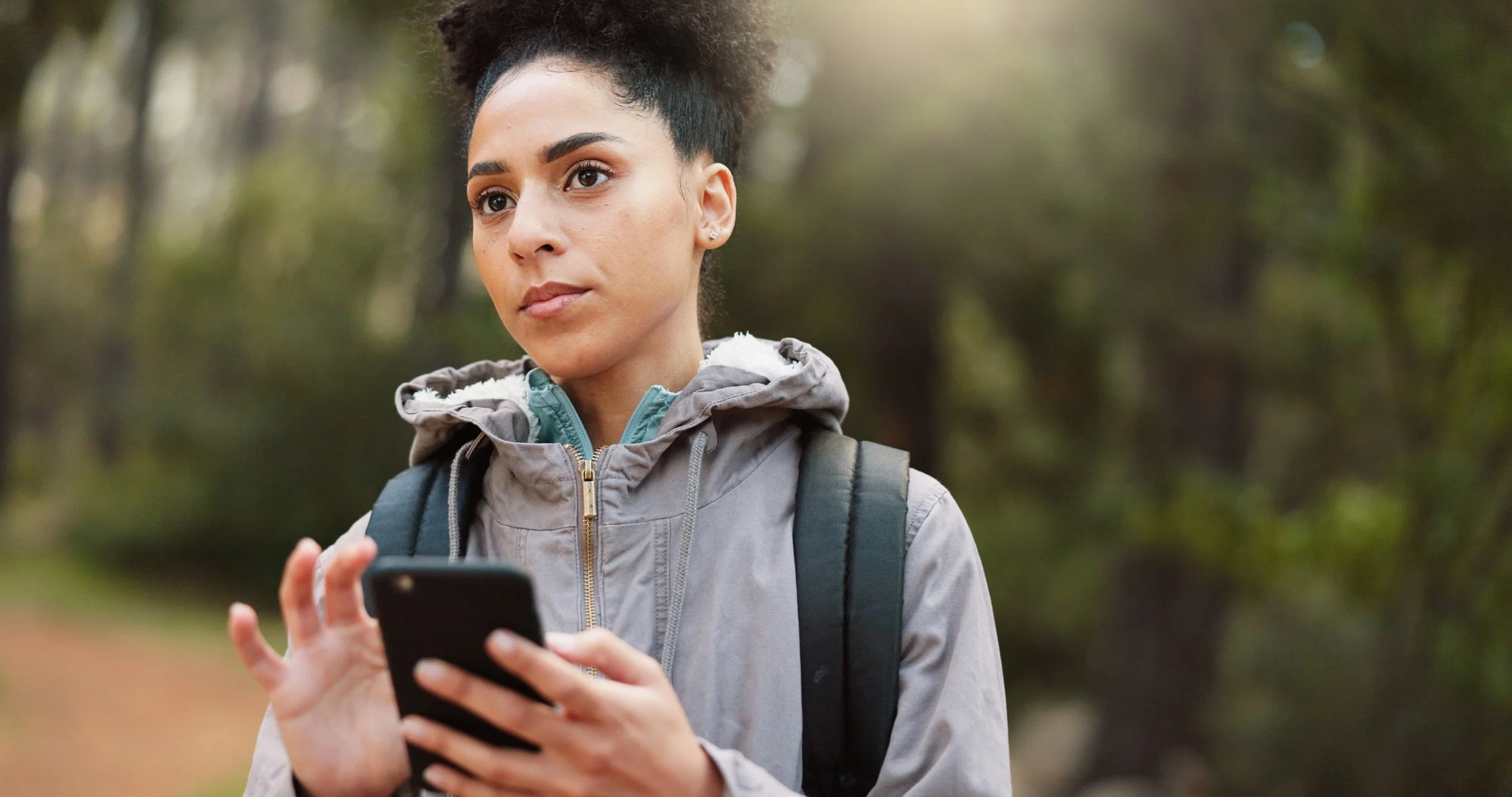 A young woman with curly hair in a ponytail is standing outdoors in a forested area, holding a smartphone and looking at it with a focused expression. She is wearing a gray hooded jacket with a backpack.