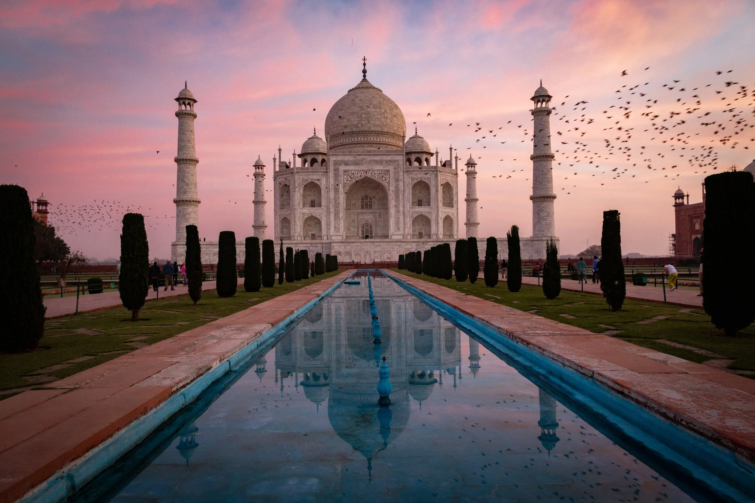 The Taj Mahal at sunset with pink and purple sky, reflecting in a long water pool, surrounded by greenery and visitors walking in the gardens.