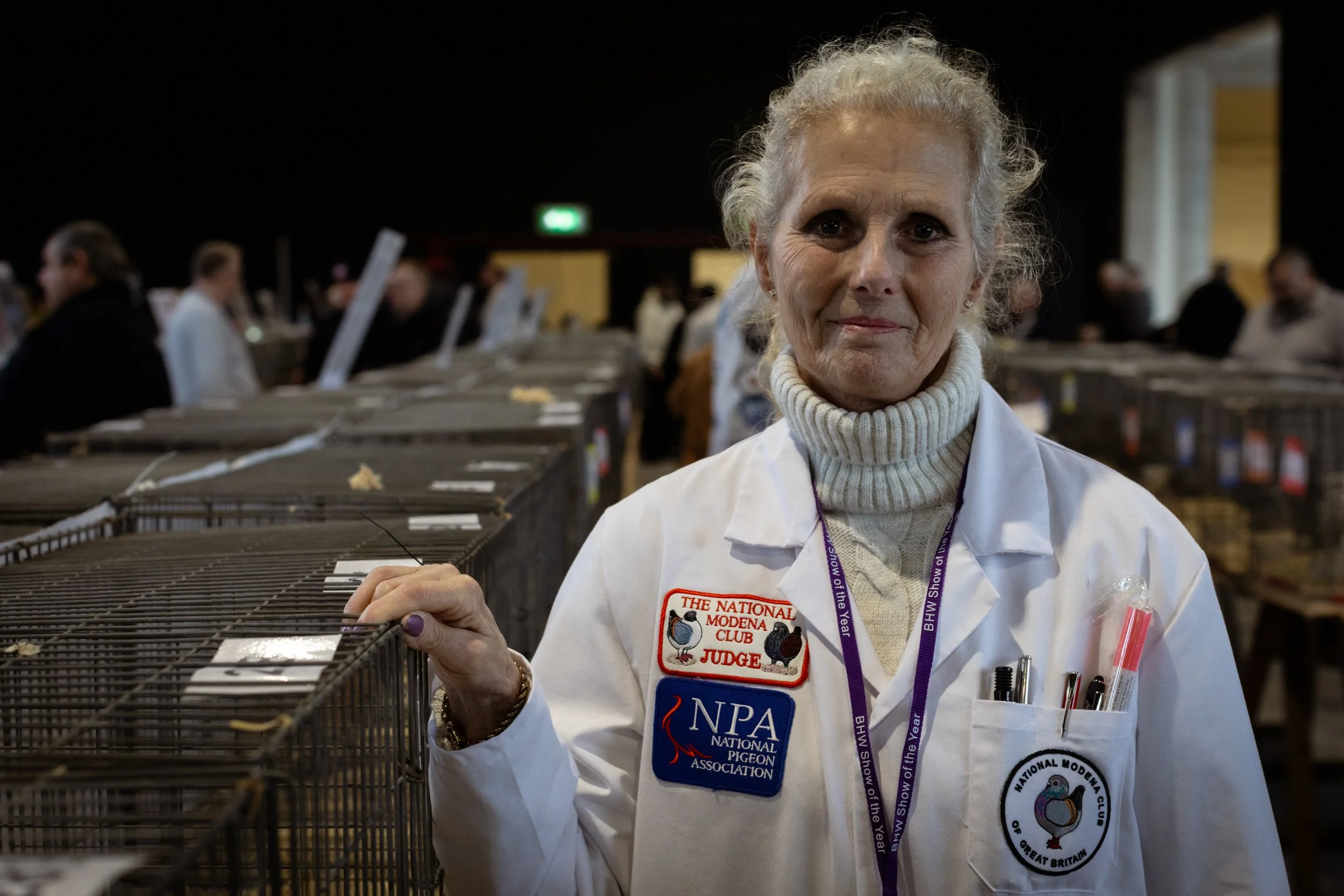 An elderly woman in a white coat with patches and pens, standing in a room with bird cages and other people in the background, at a bird judging event.