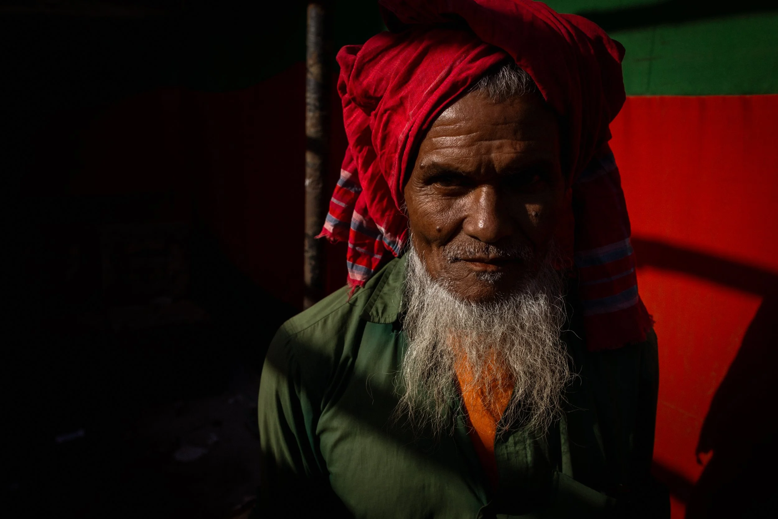 An older man with a long gray beard wearing a red turban and green shirt, sitting in shadow with bright sunlight on his face.