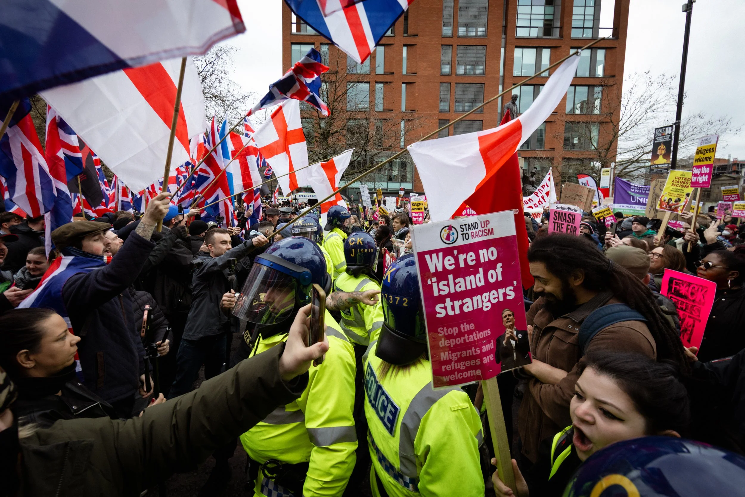 Crowd of protesters holding flags and signs, with police officers in yellow jackets and helmets, in a city street. Signs include phrases like "We're no island of strangers" and others protesting against racism and immigration policies.