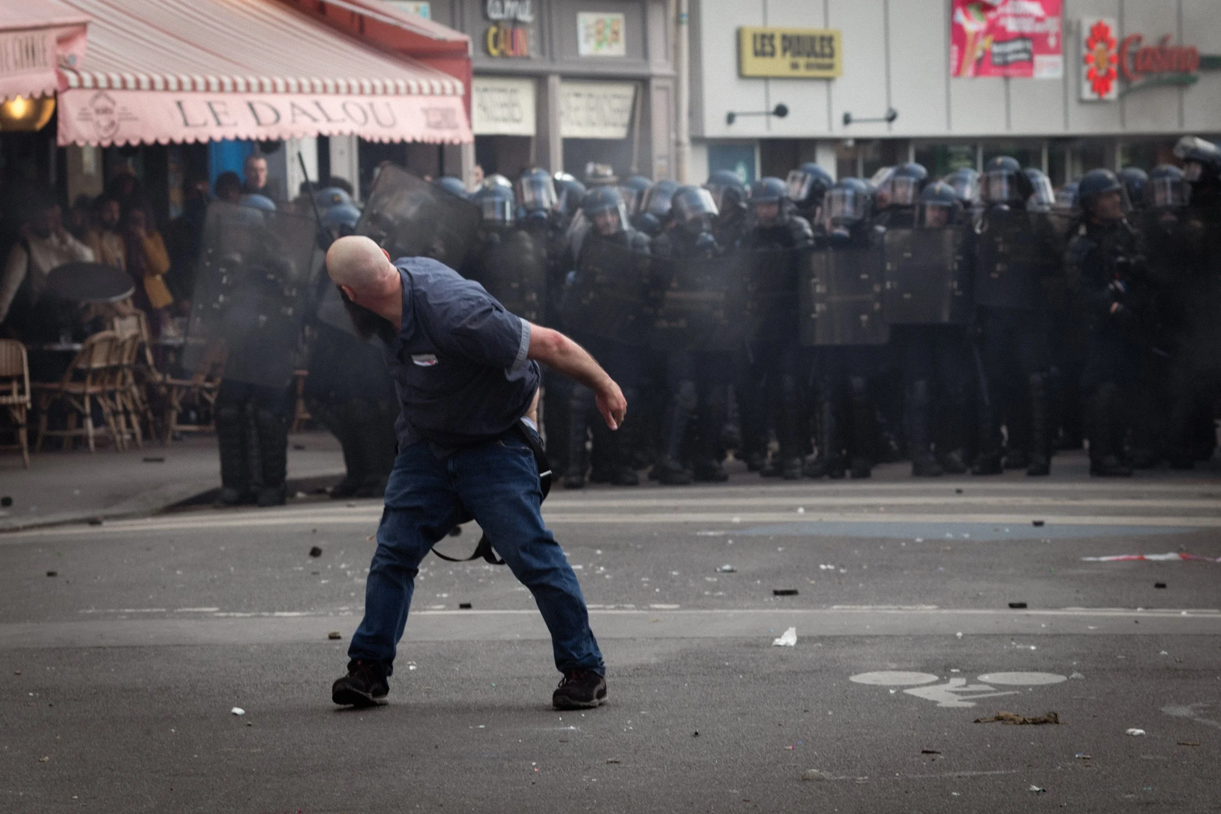 A man with a shaved head wearing a blue shirt and jeans is leaning backward after being hit or pushed, with a line of riot police in shields and helmets in the background, and a restaurant with a pink awning on the left.