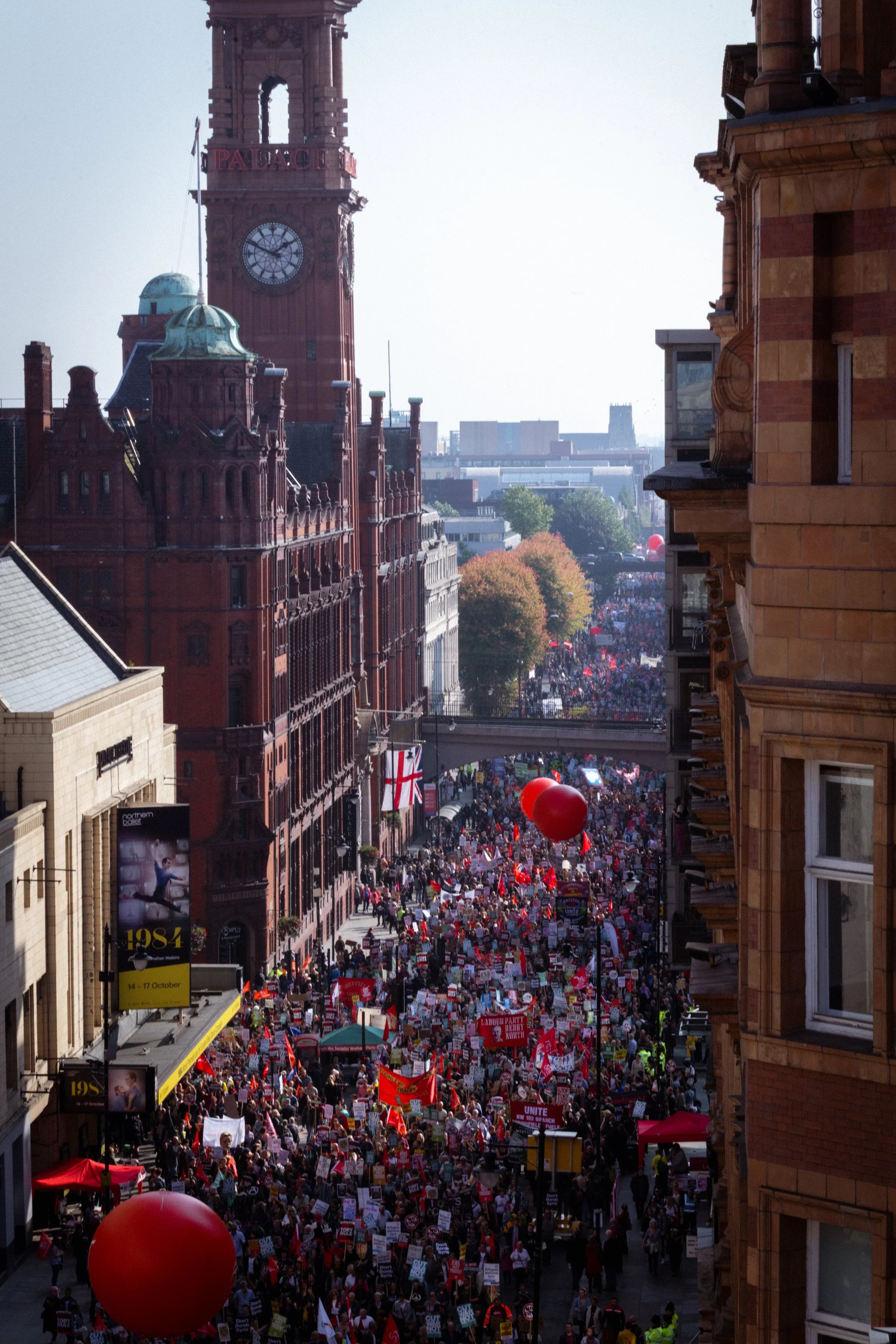 A large crowd of protesters lined up on a city street, holding signs and banners. The street is filled with people marching, with red balloons floating above the crowd. Surrounding buildings are tall and historic, with a clock tower visible in the background.