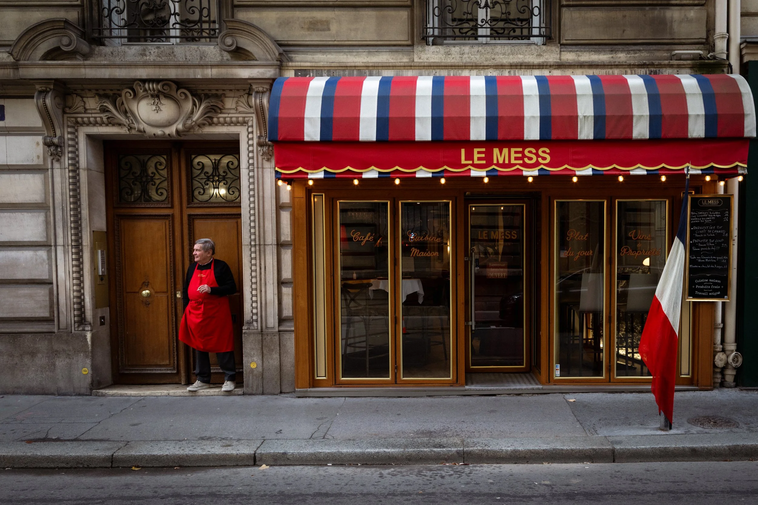 A French restaurant with a red, white, and blue striped awning, a man wearing a red apron standing outside near a wooden door, and a flagpole with the French flag.